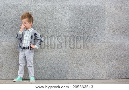 Fashion baby boy in gray jacket and mint sneakers stands on a gray wall background. Trendy boy with glasses standing on the street.