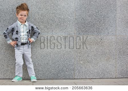 Fashion baby boy in gray jacket and mint sneakers stands on a gray wall background. Trendy boy with glasses standing on the street.
