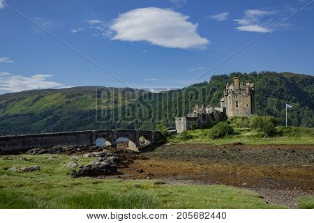 Full footbridge and Scottish Flag waving during low tide at Eilean Donan Castle on a summer's day.