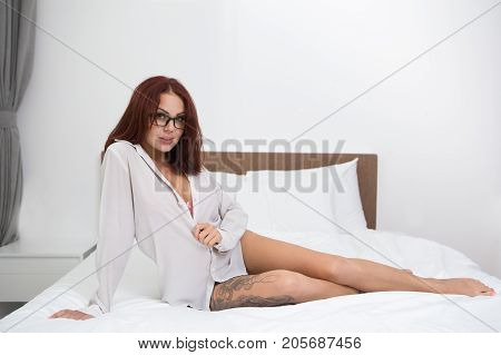 Portrait of sexy flirty tattooed young Caucasian woman wearing shirt and glasses sitting on bed, looking at camera and smiling