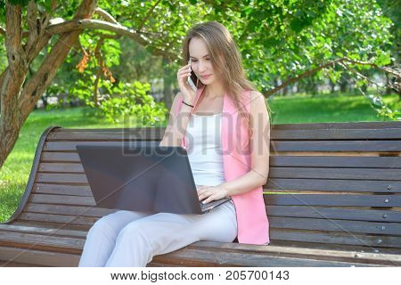 Woman sitting on bench in park talking on cell phone and using laptop