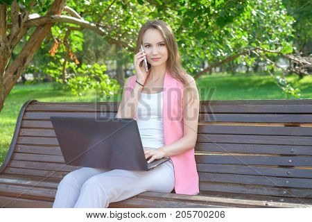 Woman sitting on bench in park talking on cell phone and using laptop