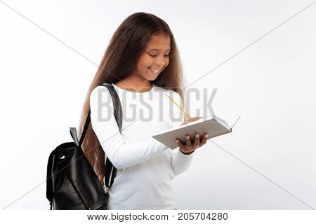 Love studying. Pretty pleasant schoolgirl making notes in her notebook while posing on a white background with a black pack on her shoulder