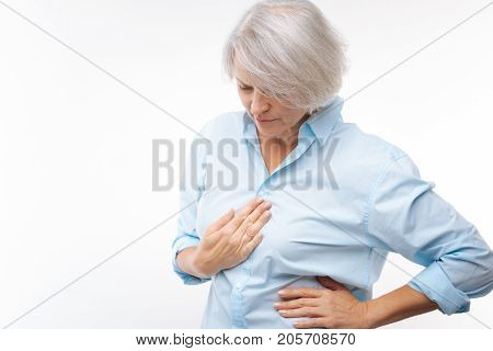 Acute pain. Middle-aged grey-haired woman in a baby blue shirt pressing her hand to the chest while suffering from stomachache
