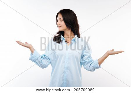 Making choice. Petite young woman in a baby blue shirt spreading her hands as if weighing decisions and making choices while looking leftwards