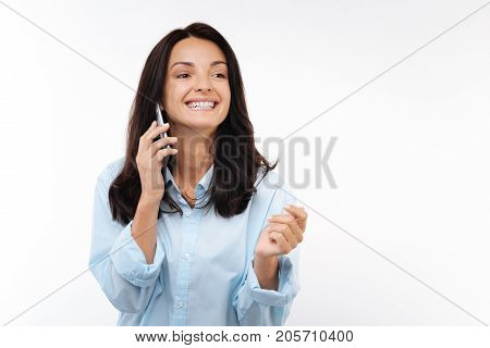 Mood-boosting chat. Beautiful dark-haired young woman talking on the phone with her friend and grinning happily while standing isolated on a white background