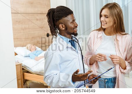 Young doctor with clipboard talking to a smiling woman in a hospital room
