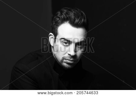 Black and white portrait of a serious young handsome man dreassed in black, dramatic lighting, looking at the camera, against plain black studio background.
