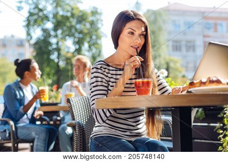 Favorite healthy drink. Charming young woman sipping smoothie with a straw and using her laptop while sitting on a terrace of a cafe