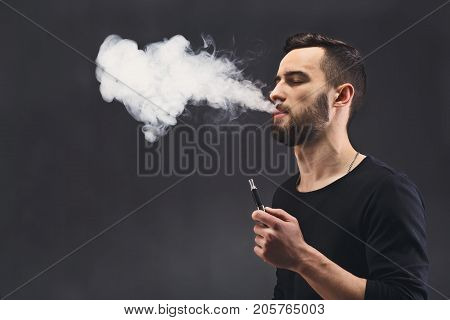Young man vaping, studio shot. Bearded guy blowing a heart-shape cloud of smoke on black background. Nicotine free smoking and vapor concept, copy space.
