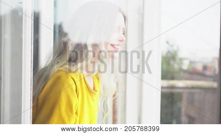 Attractive young woman in a yellow dress peeks into an open window and smiles. View from behind the glass.
