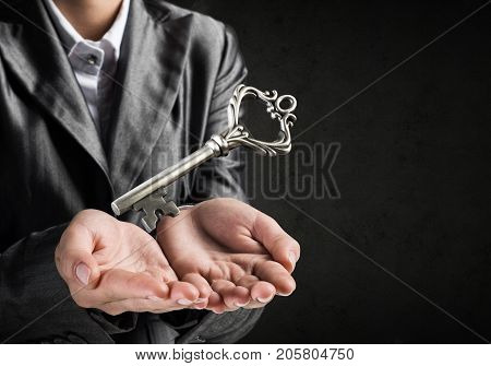 Cropped image of businessman in suit keeping big key in hands with dark wall on background.