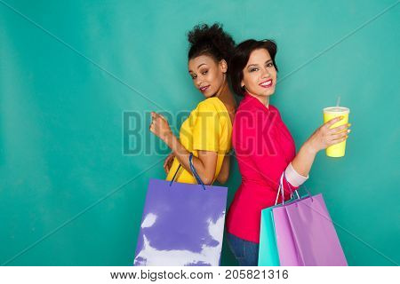 Happy multiethnic girlfriends with shopping bags and take away drinks. Two excited shopaholics at turquoise sudio background with copy space