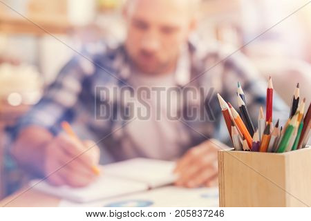 Creative workspace. Selective focus on a wooden jar with multi colored pencils standing on a table while a serious self employed man writing something down while working in the background.