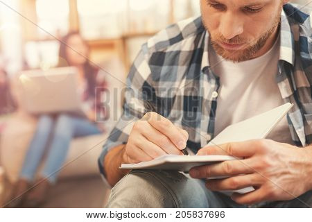 Focusing on work. Selective focus on a millennial man sitting with a notebook on his knees and writing something down while a young brunette with a laptop looking into vacancy in the background.