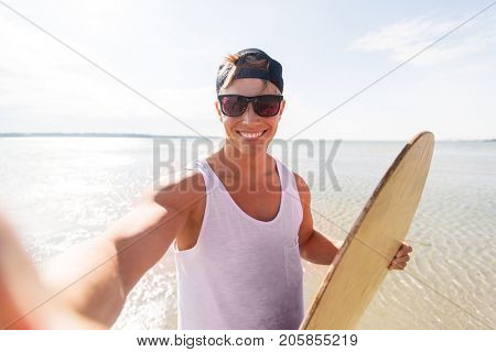 skimboarding, water sport and people concept - happy smiling young man with skimboard taking selfie on summer beach