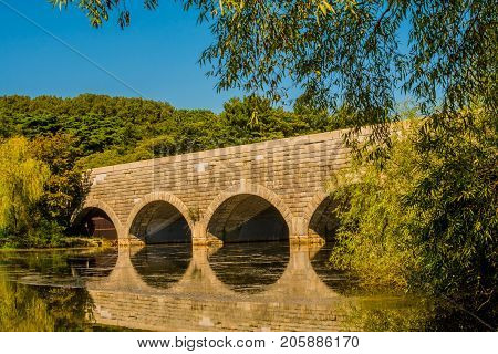 Large brick bridge with arched tunnels spanning a river with its reflection in the water surrounded by lush green trees with a clear blue sky.
