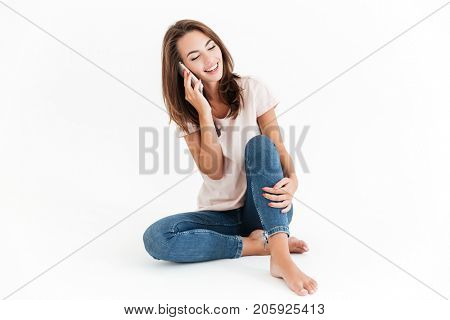 Happy brunette woman sitting on the floor and talking by the smartphone over white background