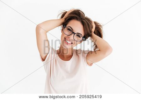 Smiling brunette woman in eyeglasses holding hair and looking at the camera over white background