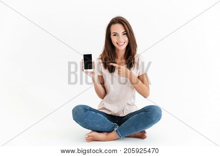 Smiling brunette woman showing blank smartphone screen and pointing at him while sitting on the floor over white background