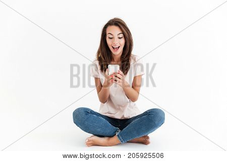Happy surprised woman sitting on the floor and writing message on smartphone over white background
