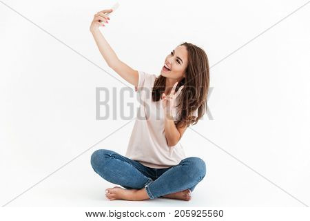Happy brunette woman making selfie and showing peace sign at camera while sitting on the floor over white background