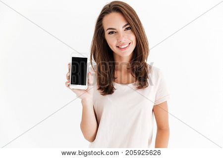 Joyful brunette woman showing blank smartphone screen and looking at the camera over white background