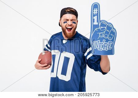 Photo of excited man fan in blue t-shirt standing isolated over white background. Looking camera wearing number one fan glove holding rugby ball.