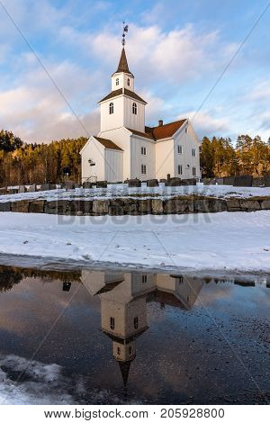 Church in march, winter landscape with snow and blue sky in Iveland in the south part of Norway. Reflections of the church in water, vertical photo