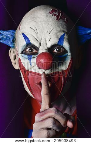 a scary evil clown peering out from a purple stage curtain, with his forefinger in front of his lips, asking for silence