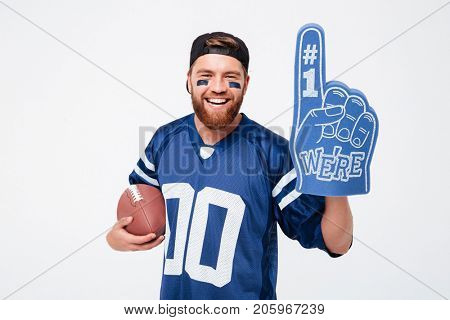 Photo of excited man fan in blue t-shirt standing isolated over white background. Looking camera wearing number one fan glove holding rugby ball.