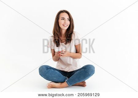 Pleased brunette woman sitting on the floor with smartphone in hands and looking away