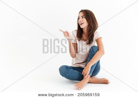 Happy brunette woman sitting on the floor and pointing away over white background