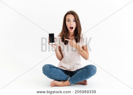 Shocked brunette woman showing blank smartphone screen and pointing at him while sitting on the floor over white background