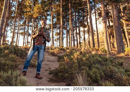 Portrait of a smiling young African man wearing a backpack standing on a trail while hiking alone in the forest on a sunny day