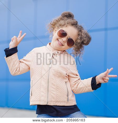 Little fashion hipster curly smiling girl in sunglasses showing peace by fingers against blye wall. Outdoor