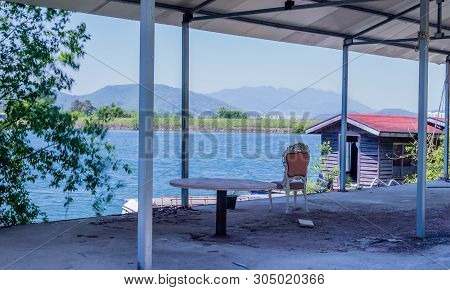 Round Table And Chair Sitting Under Metal Shelter At Old Dirty Abandoned Fishing Resort With Lake In