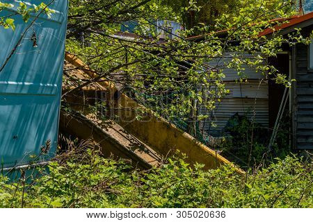 Old Yellow Fishing Boat Resting On Embankment Among Tree Branches And Bushes Behind Old Abandoned Wo