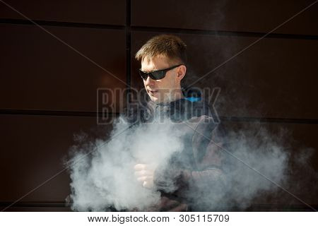 Vape Man. An Adult Bearded Man In Sunglasses Smokes An Electronic Cigarette On A Sunny Day Outside.