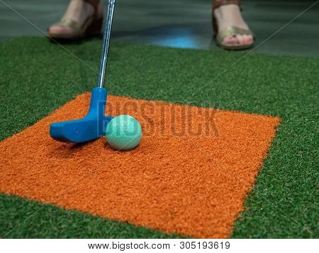 Blue Putter On Turf Next To Green Golf Ball On Miniature Golf Course With A Woman Hitting