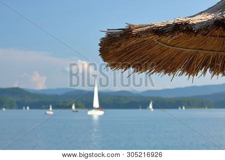 Sailing On Lake Surrounded By Hills, Yachts And Boats, Beach Straw Beach Ambrella In Foreground