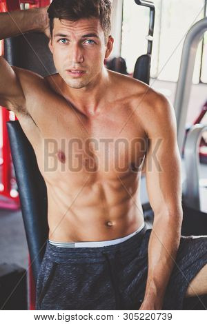 Portrait Of Very Handsome Fit Young Man With Nacked Muscular Torso Is Sitting In Gym. Natural Light