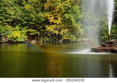 Water Jet Fountain On Rock In Center Of Lake With Colorful Fall Trees Reflected In Calm Water Surfac