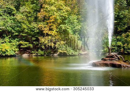 Bronze Snake Fountain On Rock In Center Of Lake With Colorful Fall Trees. Rainbow In Water Drops Fro