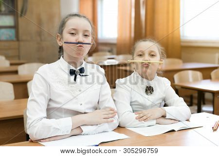 Children Indulge In School In The Classroom Make Face Mimicry. Two Schoolgirls Pinch A Pencil With T