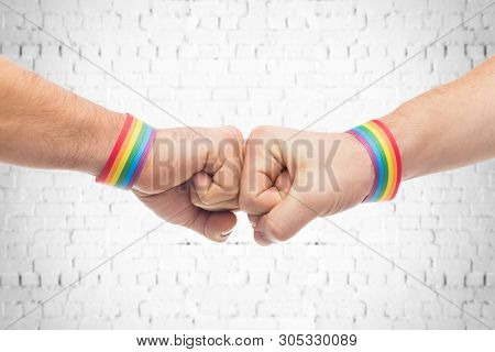 lgbt, same-sex love and homosexual relationships concept - close up of male couple hands with gay pride rainbow awareness wristbands making fist bump gesture over brick wall background
