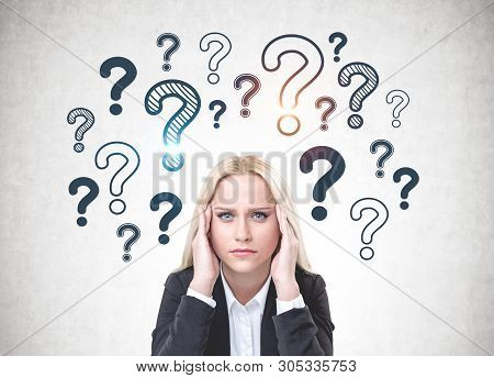 Stressed Blonde Woman In Suit Sitting Near Concrete Wall With Question Marks Drawn On It. Concept Of