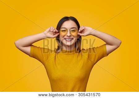 Young Lady In Trendy Yellow T-shirt Adjusting Sunglasses And Smiling For Camera Against Vivid Backgr