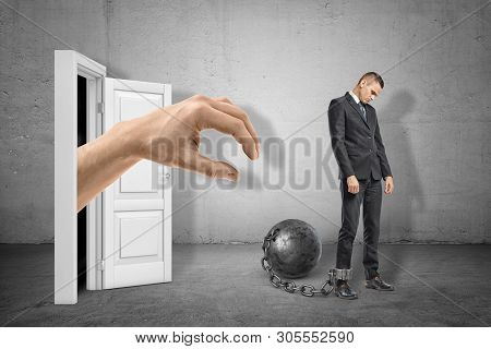 Big Hand Reaching Through White Doorway To Catch Young Sad Businessman Who Is Chained To Metal Ball 