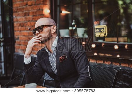 Pensive Modern Man Is Smoking A Cigarette Outside Of Coffeshop While Drinking His Cup Of Coffee.